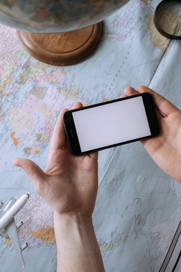 Hands holding a smartphone with blank screen above a world map, showcasing global travel planning concept.