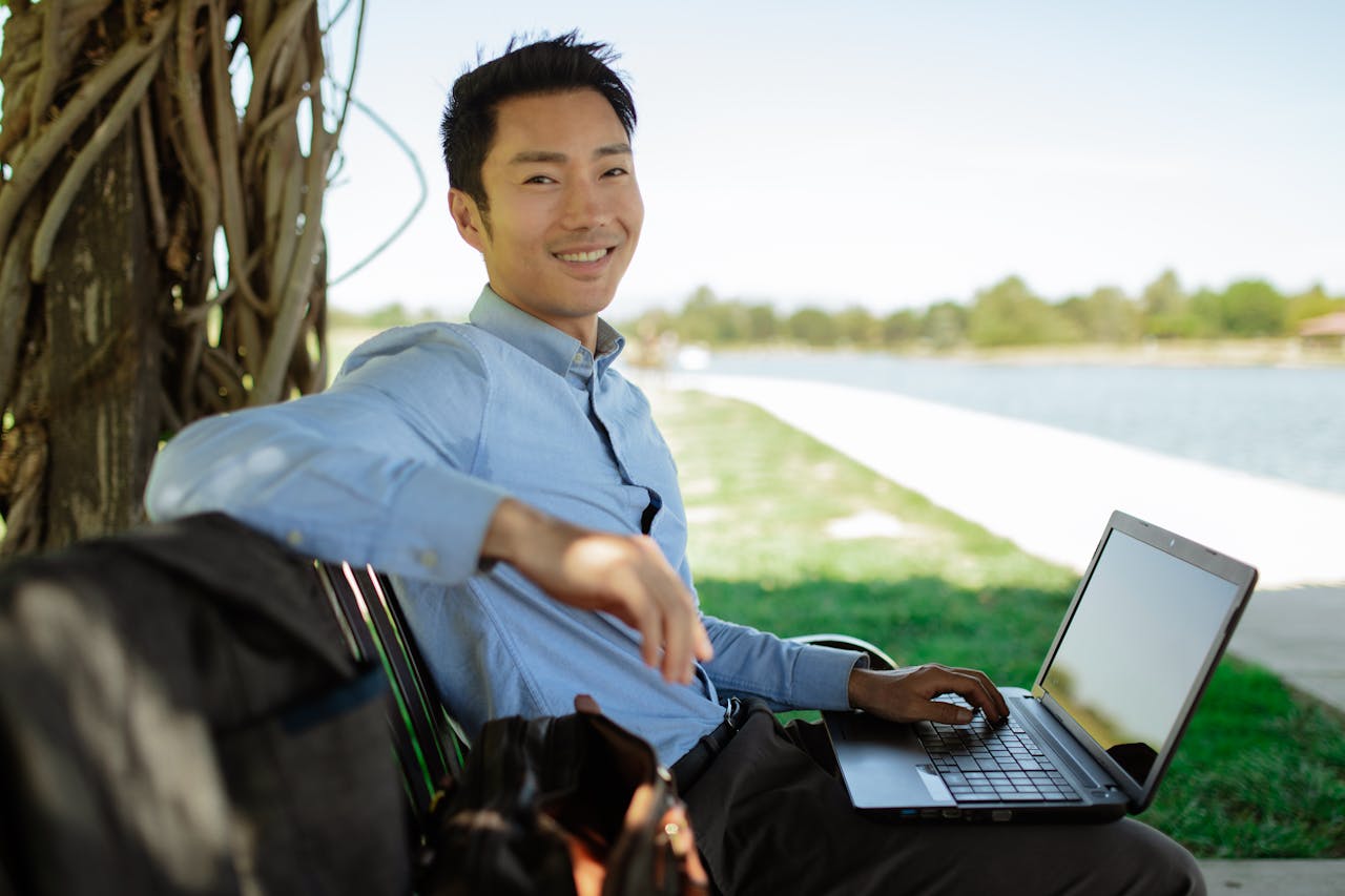 Asian man in a park sitting on a bench, using a laptop, smiling at the camera.