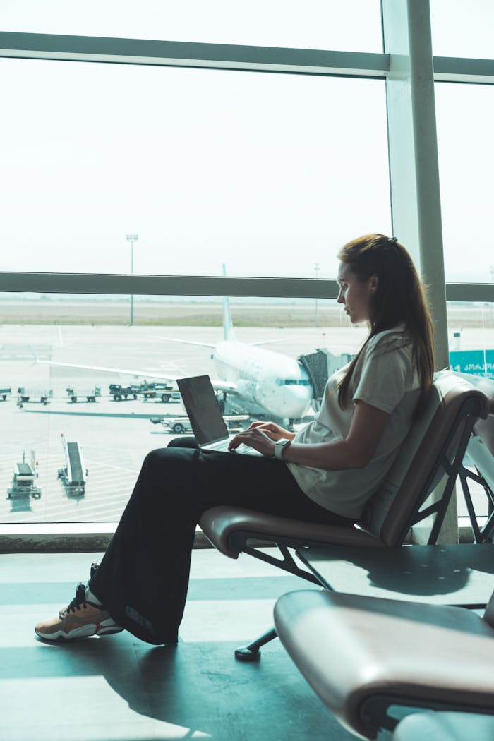 services-03 Side view of woman using laptop at airport, airplane visible outside through large glass windows.